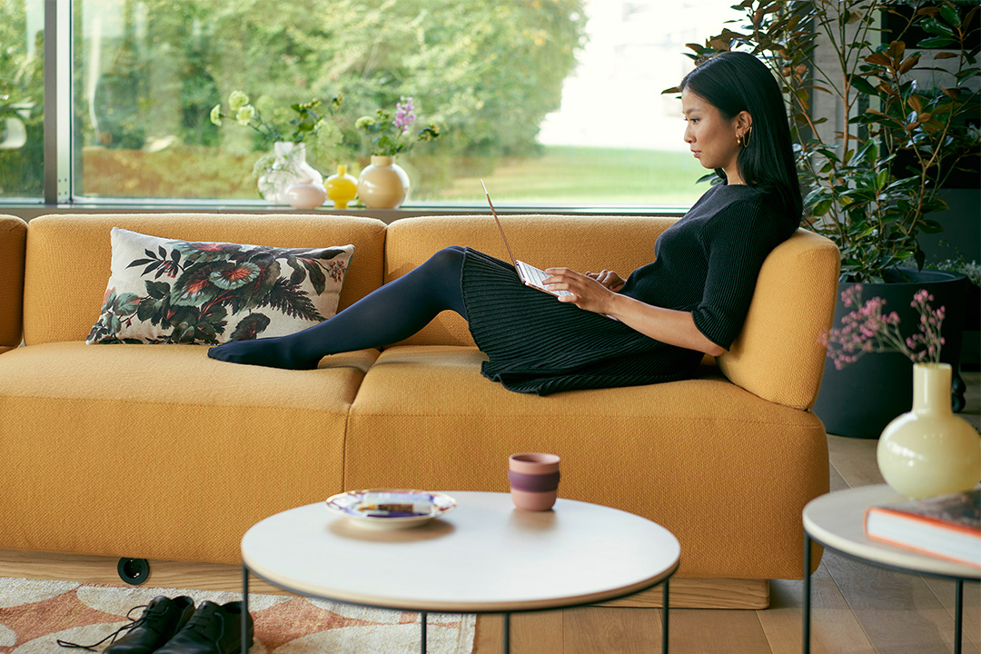 Eine Frau sitzt entspannt auf einem se:living von Sedus und arbeitet mit einem Laptop, der auf ihren Oberschenkeln ruht. Im Vordergrund ist unscharf ein se:living side table zu erkennen, der die gemütliche und flexible Atmosphäre des Raumes unterstreicht. Im Hintergrund dominiert ein großes Fenster, das den Raum mit natürlichem Licht erfüllt und einen weiten Blick auf den Garten bietet. Diese Szene veranschaulicht das Konzept des duktilem Raumes, der als flexibler Knotenpunkt in hybriden Büros dient und individuelles Arbeiten sowie den Übergang zu dezentralen Arbeitsmethoden unterstützt.