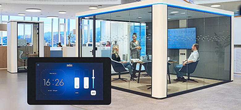 Moderno spazio ufficio con una sala riunioni con pareti in vetro dove sono sedute tre persone e una è in piedi, con un display touch digitale che mostra l’ora e i comandi in primo piano.