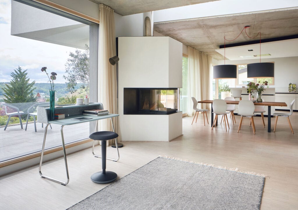 Eine moderne Wohn- und Arbeitsumgebung mit großen Glasfenstern, die einen Panoramablick auf die Natur bieten. Der Innenraum ist minimalistisch gestaltet, mit einem eleganten Kamin im Zentrum, einem Schreibtisch in der Nähe der Fensterfront und einem Esstisch mit modernen Stühlen im Hintergrund. Große Pendelleuchten verleihen dem Raum eine angenehme Atmosphäre. Ideal für SEO-Schlüsselwörter wie "modernes Interieur", "Panoramablick", "Wohn- und Arbeitsbereich" und "minimalistisches Design".